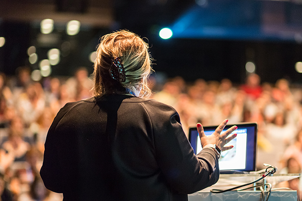 Female speaker giving a talk on corporate business conference. Unrecognizable people in audience at conference hall. Business and Entrepreneurship event.