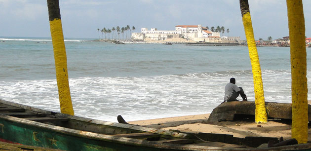 A man sitting on a boat on the beach with the Cape Coast Castle in the Background - Unexplored West Africa
